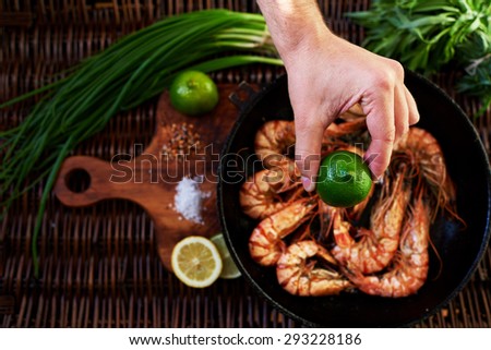 In the foreground, a man\'s hand squeezes a lemon on fried tiger prawns, situated near the green onion bunch of rosemary, In the old cast iron skovordke The royal fried shrimp