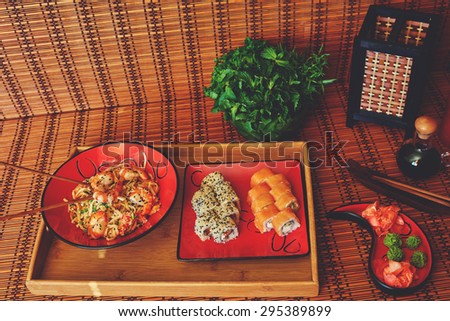 On a wooden tray, there are two red plates on a single two servings of sushi with salmon and Philadelphia cheese with smoked eel and the second plate udon noodles with  peppers onion and shrimp sauce