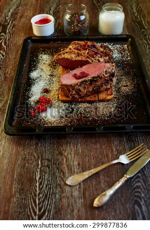 Top view on the table is a pan which is a large piece of lamb, standing beside spicy sauce with the addition of sour wild berries next to a baking tray are silver cutlery