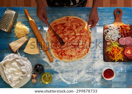 Housewife smeared a thin spicy tomato sauce pizza dough, pizza lies on textural blue table near A wooden cutting board on which there are all the ingredients chopped