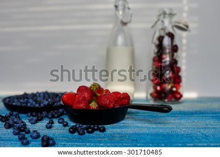 On a blue vintage wooden desk are two cast iron pans that sprinkling strawberries and blueberries near the scene along there are two glass bottles with milk and cherries