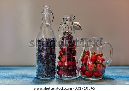 On the vintage wooden table, there are three glass carafe which are prepared fresh berries for cooking stewed fruit