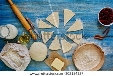 On a bright blue vintage wooden table dough is cut into triangular piece near Cherry, sieve and milk