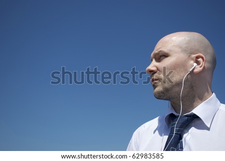 Royalty free stock image of an unshaven office worker wearing white headphones looking out to distance against clean blue sky providing copy space to the left, vignetted