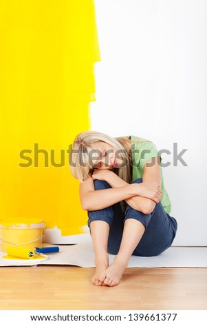 Exhausted woman taking a break from decorating her house and painting the walls sitting on the floor with her head on her arms and a half painted yellow wall behind