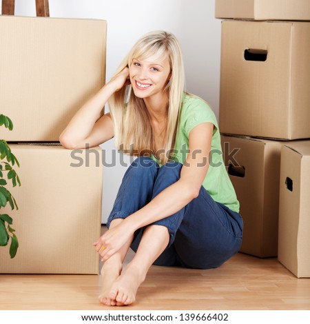 Beautiful smiling young woman sitting barefoot on the floor surrounded by cardboard cartons when packing to move house
