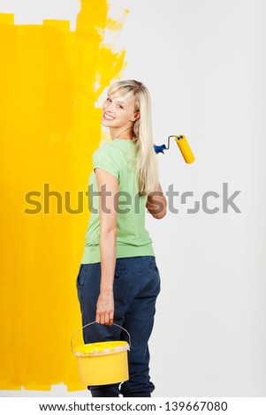 Smiling young woman carrying yellow paint in a container and a roller in her hand turning back to look at the camera as she prepares to continue decorating her house