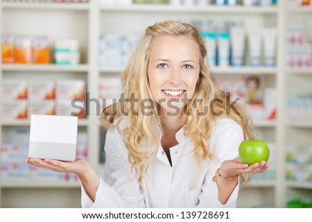 Portrait of confident female pharmacist holding pill box and an apple in pharmacy