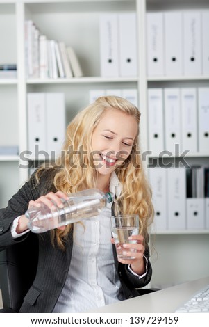 Portrait of happy young businesswoman pouring water in glass from bottle at office