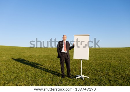 Mature businessman with hand in pocket standing by blank flipchart on grassy field against clear sky