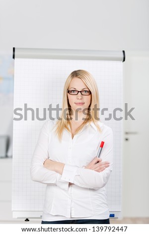Portrait of confident blond young woman standing by flip chart in office, arms crossed