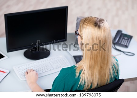 Rear view of young blond businesswoman using desktop computer in office