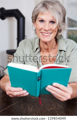 Modern attractive smiling senior lady with short grey hair reading a book sitting indoors