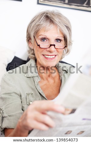 Retired woman wearing glasses sitting reading a newspaper at home on a sofa