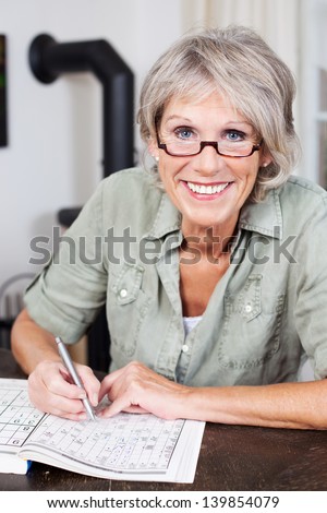Smiling Attractive Elderly Woman Wearing Glasses Sitting At A Table Doing A Crossword Puzzle In A Book