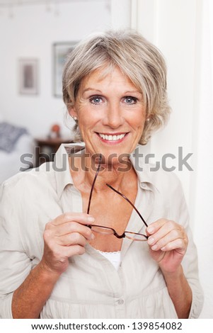 Modern Older Woman Standing In Her House Holding Her Glasses In Her Hand Smiling At The Camera