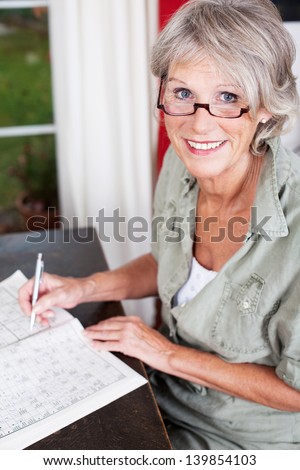 Older Woman Wearing Glasses Working On A Crossword Puzzle In A Puzzle Book Sittiing At A Small Wooden Table In Her House