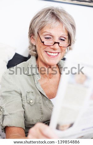 Elderly grey haired lady wearing a pair of reading glasses sitting reading the newspaper with a smile