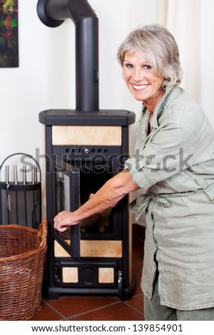 Attractive smiling senior lady puting wood in the stove or woodburner to heat her house during winter