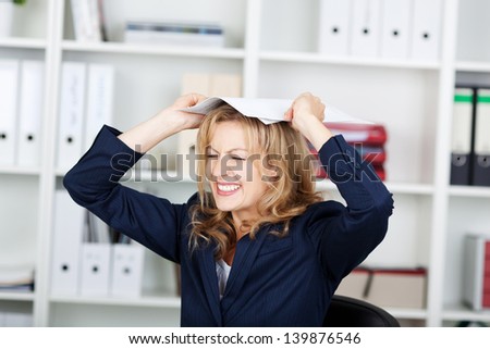 Angry businesswoman clenching teeth while placing documents on head in office
