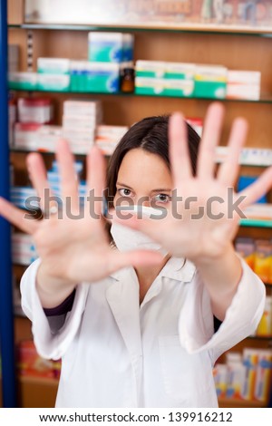 Portrait of mid adult female pharmacist in mask gesturing stop sign at pharmacy