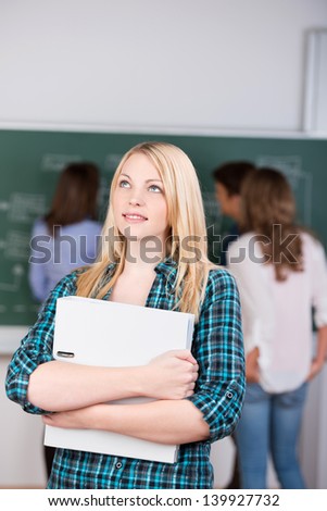 Thoughtful female student holding binder while looking up with teacher teaching students on chalkboard in background