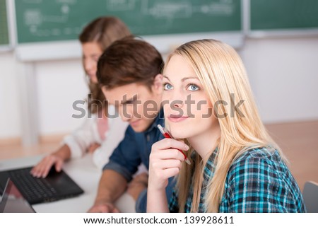 Thoughtful young female blond student looking up with classmates at classroom desk