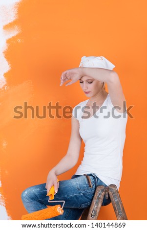 Tired young woman relaxing on ladder while holding paint roller against orange colored wall