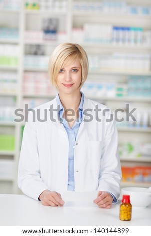Portrait of young female pharmacist with pill bottle and prescription paper standing at pharmacy counter
