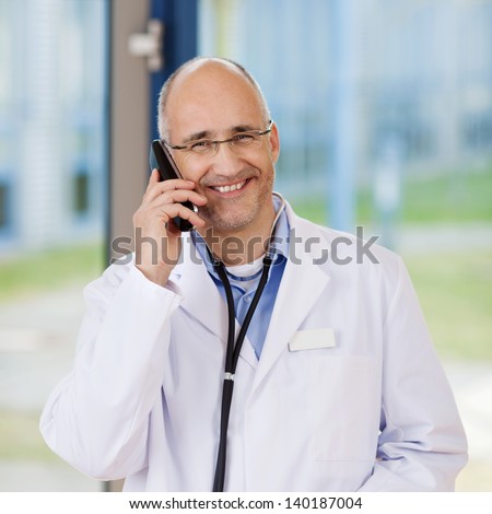Mature male smiling doctor using mobile phone in clinic
