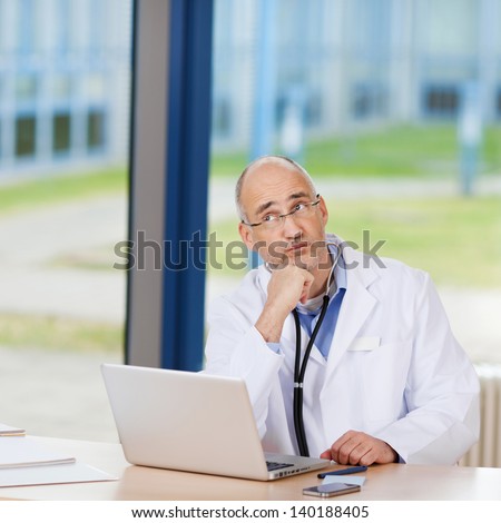 Thoughtful mature doctor with laptop on desk in clinic
