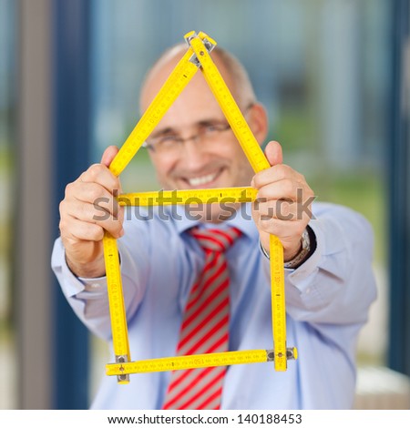 Portrait of happy mature businessman holding house made of ruler in office