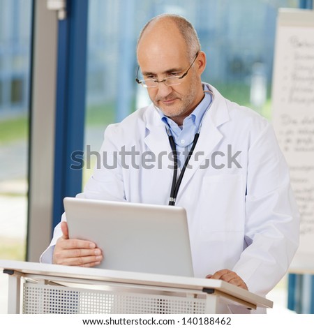 Mature male doctor using laptop at podium in clinic