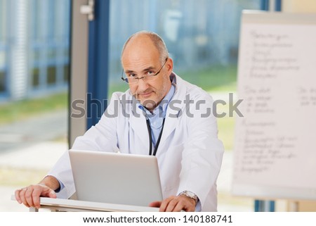 Portrait of confident mature doctor with laptop at podium in clinic