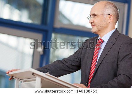 Confident mature businessman looking away while standing at podium in office