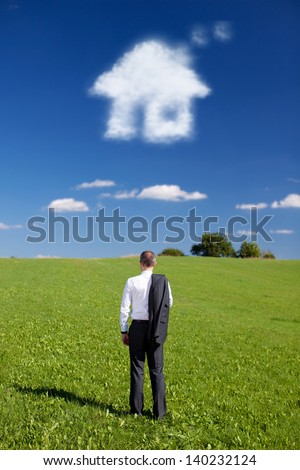 Man dreaming of a new home standing with his back to the camera and his suit jacket slung over his shoulder facing out over a green field with a cloud in the shape of a house in the blue sky