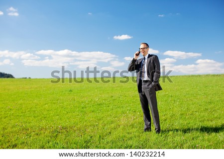 Businessman standing in his suit in a green sunny field under a blue sky phoning from his mobile phone with copyspace