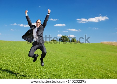 Excited businessman jumps high in the air cheering and celebrating a success or achievement in a green sunny field under a blue sky
