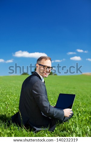 A handsome young businessman working out in nature sits on the grass in a green field working on his laptop and looking back at the camera