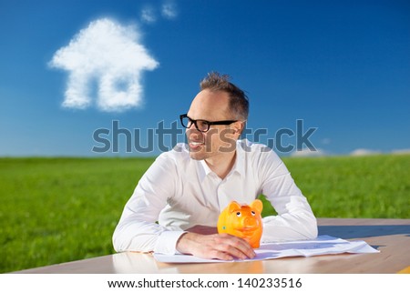 Happy man with his piggy bank sitting at a table in a green field under a sunny blue sky dreaming of a new home with a cloud in the shape of a house above his head
