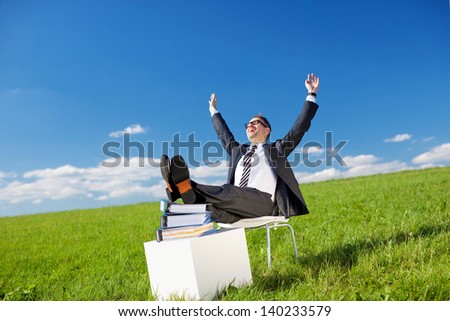 Businessman relaxing in the fresh air sitting on a chair in a green field with his feet up on a pile of documents and his arms raised to the sun
