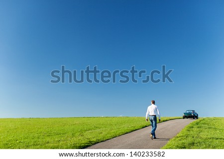 Man walking along a tarred road running through a green field towards his cabriolet car parked on the horizon against a sunny blue sky