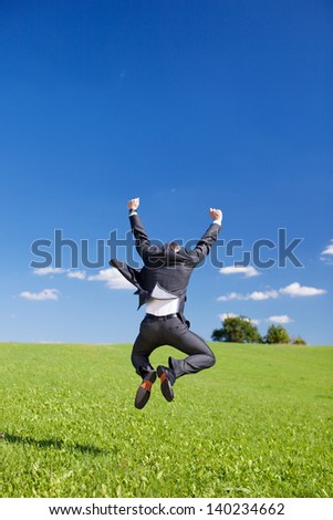 Jubilant businessman jumping for joy with his arms raised and back to the camera in a lush green field