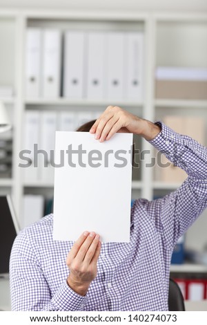 Young businessman holding blank paper in front of face in office