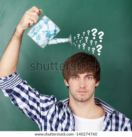 Portrait of young male student holding watering can with question marks on chalkboard representing confusion