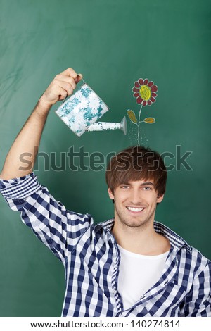 Portrait of handsome young male student holding watering can with flower drawn on chalkboard representing growth of ideas