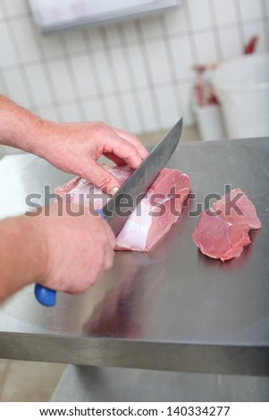 Close-up of the hands of a butcher slicing steaks with a large knife from a large piece of fresh meat over a stainless steel workbench