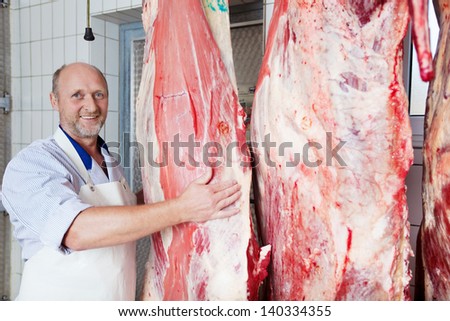 Happy middle age butcher near three cow carcass halves in the butchery