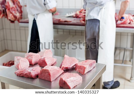 Several portions of fresh cut meat resting on a stainless steel workbench, with two butchers working on the background