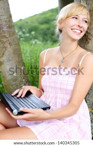 Vivacious woman using her laptop in nature sitting with her back against the trunk of a tree in lush green grass